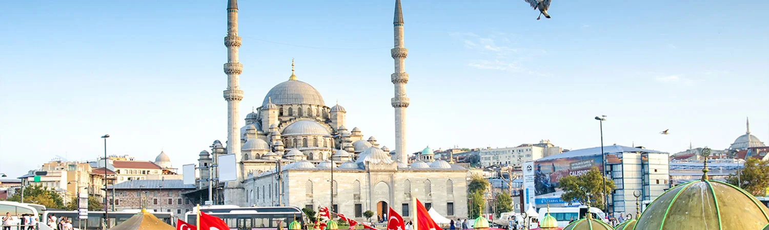 A bustling scene in front of a grand mosque with towering minarets, surrounded by buildings and tents, under a clear blue sky.