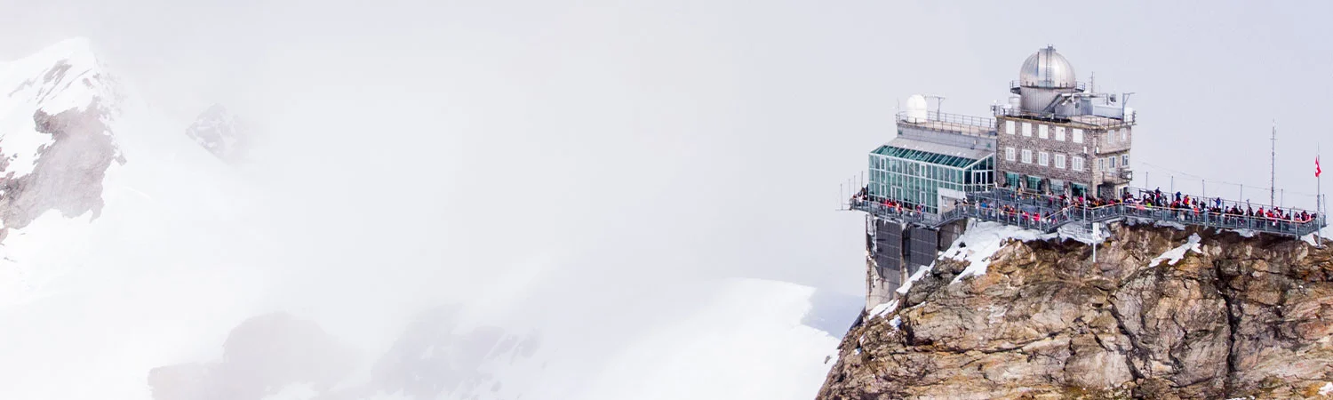 A crowded observation deck on a rocky mountain peak amidst the snowy alps, covered in mist, offering a panoramic view to tourists.