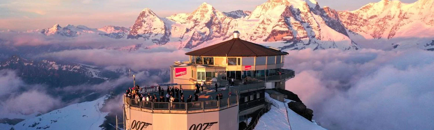 Breathtaking view of the 007 Elements museum perched on a snowy mountain peak, surrounded by majestic alps and enveloped in clouds illuminated by the golden hue of sunset.