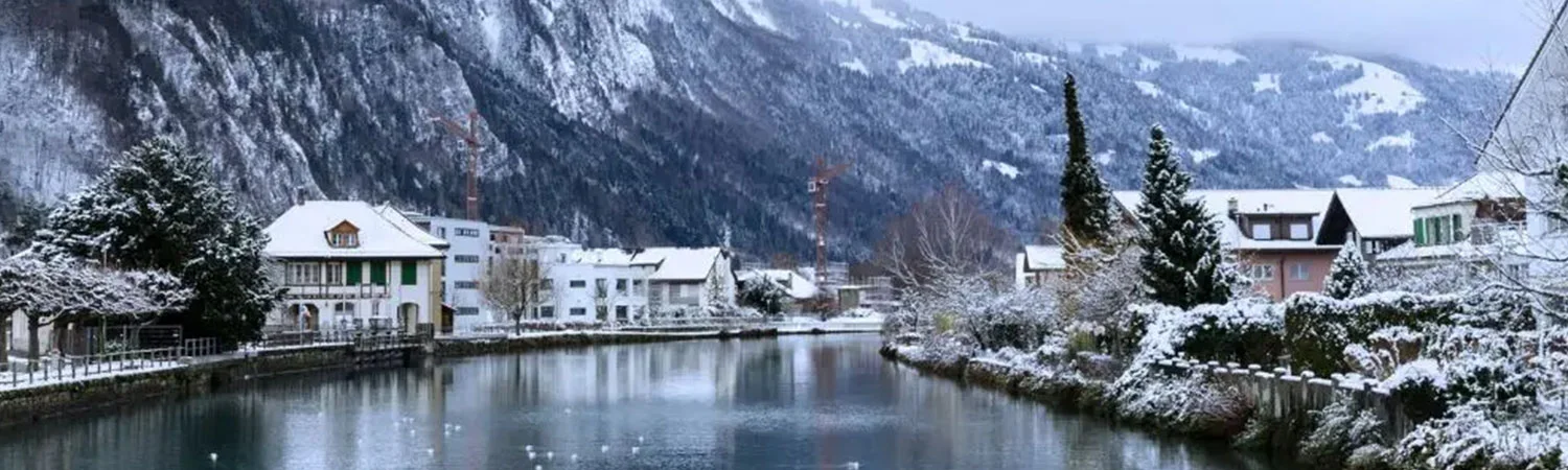 A serene winter landscape featuring a calm lake surrounded by snow-covered trees and buildings, with majestic snow-capped mountains in the background.