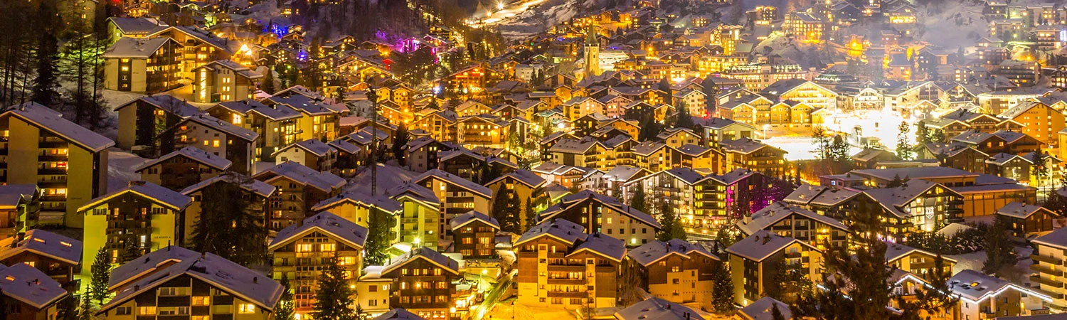 A breathtaking night view of a snow-covered mountain town, illuminated buildings casting a warm glow against the cold winter backdrop, showcasing the serene beauty 