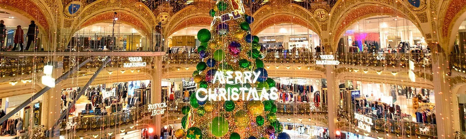 A bustling shopping mall adorned with festive Christmas decorations, including a towering, illuminated Christmas tree with a ‘Merry Christmas’ sign, surrounded by busy shoppers and an array of stores.