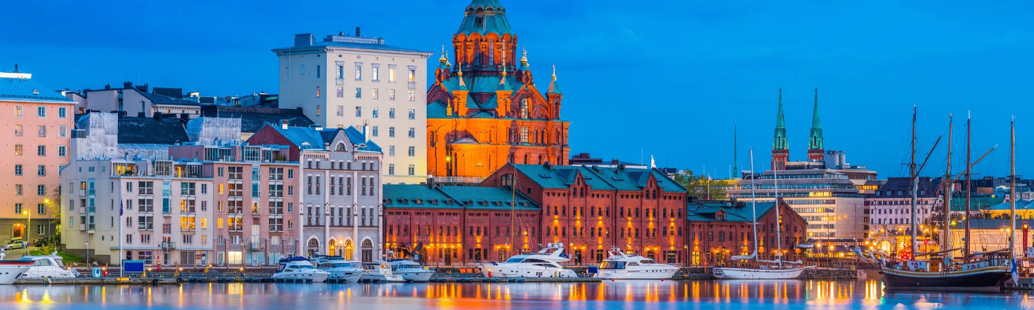 Scenic view of a harbor at dusk, with illuminated buildings reflecting on the calm water, including a prominent red cathedral, various architectural styles of buildings, and docked boats.