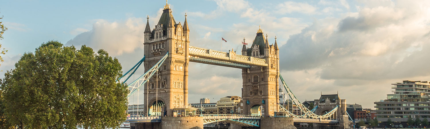 A scenic view of the iconic Tower Bridge in London, surrounded by lush green trees and modern architecture, under a bright, partly cloudy sky.