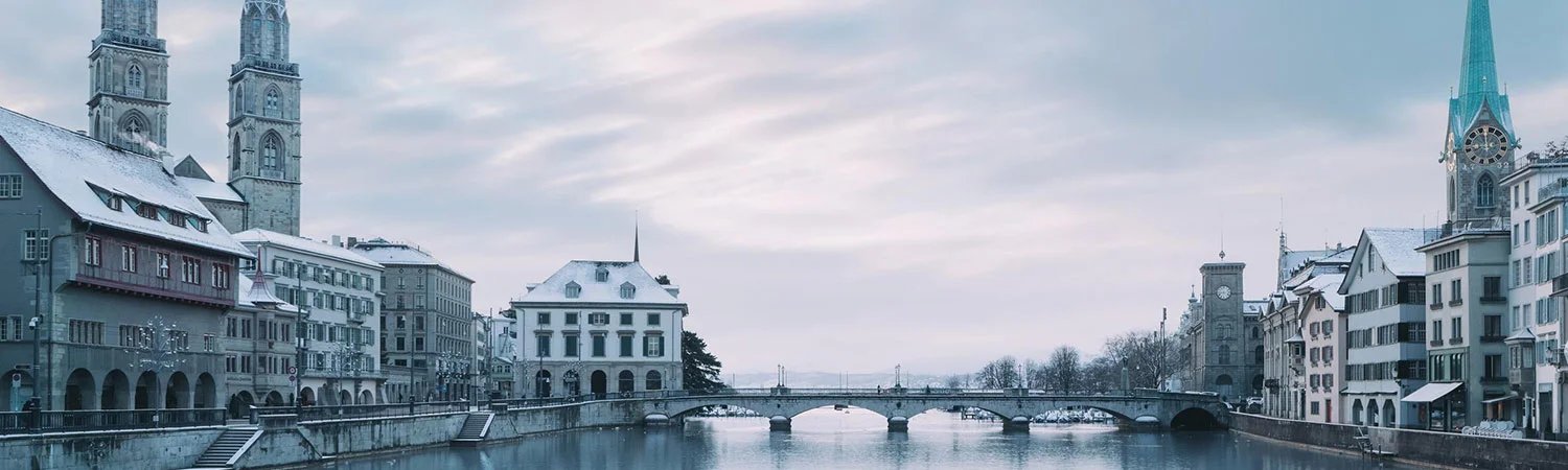 The image captures a serene view of Zurich, showcasing the iconic Grossmünster Church with its twin towers, elegant architecture along the Limmat River, and the picturesque cityscape under a cloudy sky.