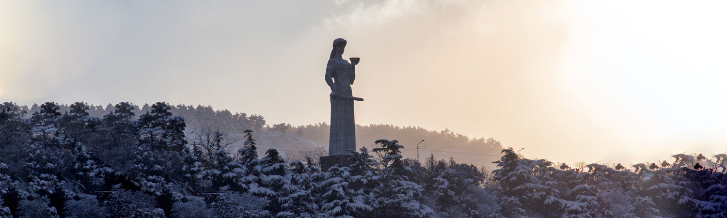 Silhouette of a statue on a column, surrounded by snow-covered trees under a bright sky in Tbilisi, Georgia