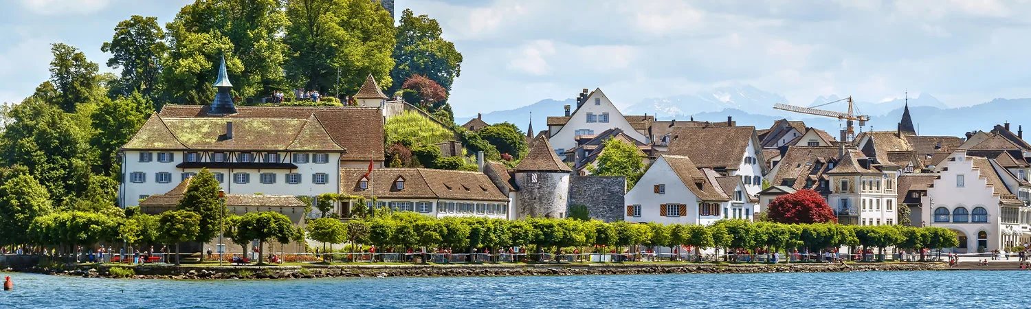 Picturesque European village by a serene lake, featuring traditional architecture, lush greenery, and mountains in the background under a clear blue sky
